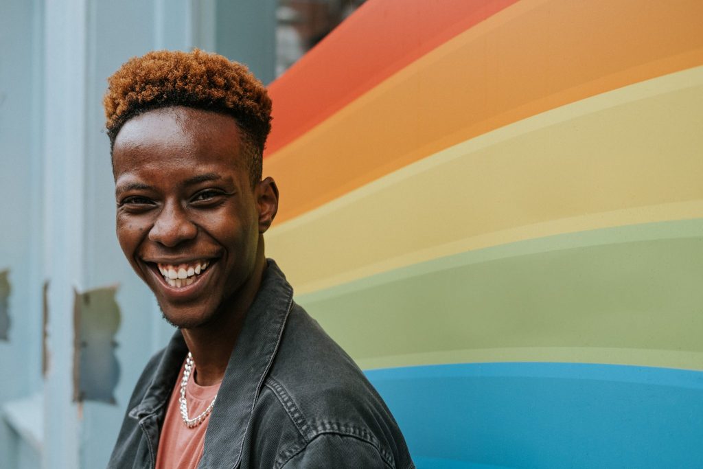 Ein junger Mann mit schwarzer Jeansjacke und dunkelbraunen kurzen Haaren steht vor der Pride Flagge, die an ein Schaufenster geklebt ist und lacht in die Kamera. ((Artikel: Was ist LGBT / LGBTQIA+ und warum wir alle so verschieden sind)