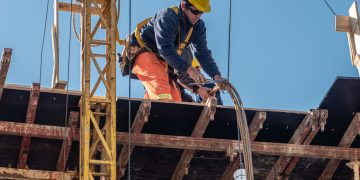 Ein Bauarbeiter mit orange-blauer Kleidung und gelbem Schutzhelm steht auf einem Dach vor blauem Himmel