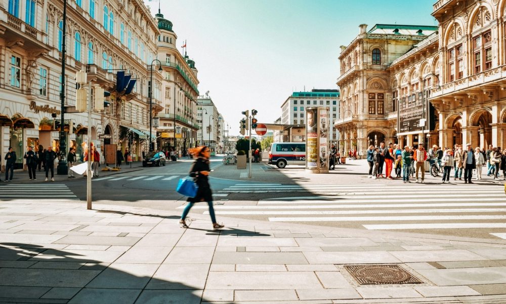 Fußgänger:innen überqueren eine Straße im Zentrum von Wien, im Hintergrund historische Gebäude, Geschäfte und die Wiener Staatsoper bei sonnigem Wetter.