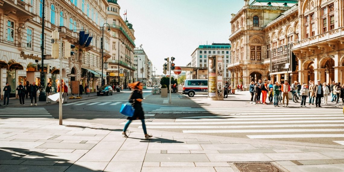 Fußgänger:innen überqueren eine Straße im Zentrum von Wien, im Hintergrund historische Gebäude, Geschäfte und die Wiener Staatsoper bei sonnigem Wetter.