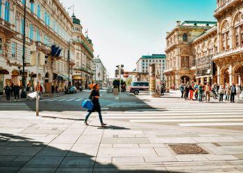 Fußgänger:innen überqueren eine Straße im Zentrum von Wien, im Hintergrund historische Gebäude, Geschäfte und die Wiener Staatsoper bei sonnigem Wetter.