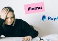 Frau sitzt am Tisch und hält sich mit genervtem Gesichtsausdruck den Kopf, vor ihr liegen Rechnungen und Papierkram. Logos von Klarna und PayPal