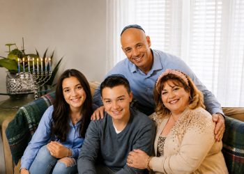 Eine jüdische Familie sitzt lächelnd gemeinsam im Wohnzimmer; die Kippas der beiden männlichen Familienmitglieder und der Chanukka-Leuchter im Hintergrund machen jüdisches Leben sichtbar. Das Bild ergänzt den Beitrag „Was ist Antisemitismus?“ und steht für die Auseinandersetzung mit Antisemitismus heute, antisemitischen Vorurteilen und der Ideologie hinter Judenfeindlichkeit.