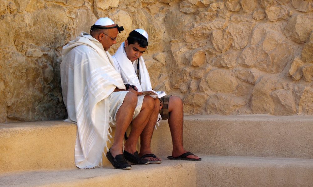 Zwei jüdische Männer mit Kippa, Tallit und Tefillin lesen gemeinsam aus einem Gebetbuch. Das Bild zeigt jüdisches Leben und begleitet den Beitrag „Was ist Antisemitismus?“. Darin wird Antisemitismus erklärt – von seiner Ideologie und Geschichte bis zu Antisemitismus heute und antisemitischen Vorurteilen.