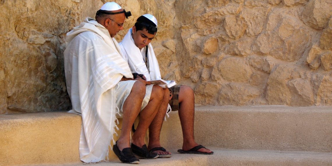 Zwei jüdische Männer mit Kippa, Tallit und Tefillin lesen gemeinsam aus einem Gebetbuch. Das Bild zeigt jüdisches Leben und begleitet den Beitrag „Was ist Antisemitismus?“. Darin wird Antisemitismus erklärt – von seiner Ideologie und Geschichte bis zu Antisemitismus heute und antisemitischen Vorurteilen.