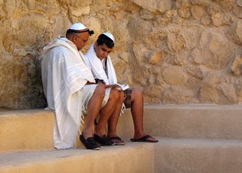 Zwei jüdische Männer mit Kippa, Tallit und Tefillin lesen gemeinsam aus einem Gebetbuch. Das Bild zeigt jüdisches Leben und begleitet den Beitrag „Was ist Antisemitismus?“. Darin wird Antisemitismus erklärt – von seiner Ideologie und Geschichte bis zu Antisemitismus heute und antisemitischen Vorurteilen.